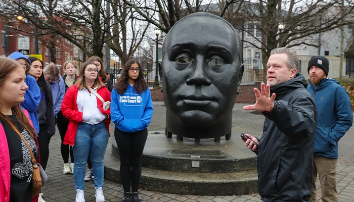 an instructor stands in front of a sculpture and speaks to students