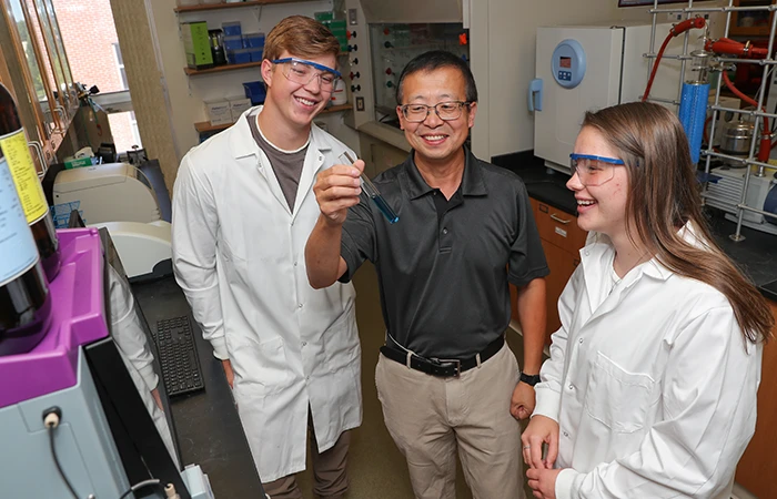 an instructor holds up a vial, showing it's contents to two students in lab coats