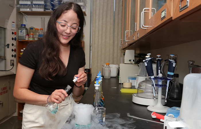 a student in a lab pours solution into a glass that is pouring out fog 