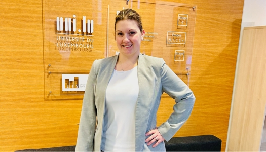 Woman in white shirt with a gray blazer standing in front of a sign that reads, "Universite Du Luxembourg."