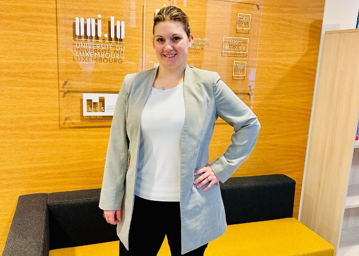 Woman in white shirt with a gray blazer standing in front of a sign that reads, "Universite Du Luxembourg."