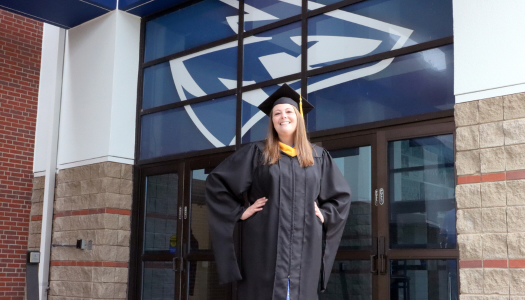 Woman with long brown hair stands with her hands on her hips in a graduation cap and gown in front of doors with a Loper head above it.