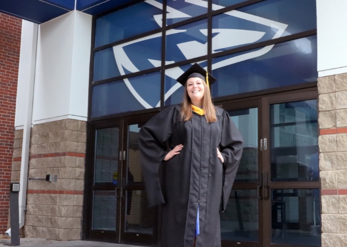 Woman with long brown hair stands with her hands on her hips in a graduation cap and gown in front of doors with a Loper head above it.