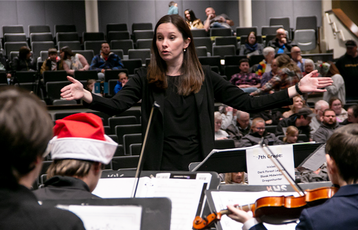 A conductor directs a 7th-grade orchestra performance, with an audience observing in a concert hall.
