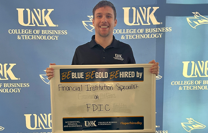 Logan Shaw holding a sign that reads, "Be Blue. Be Gold. Be Hired by Financial Institution Specialist at FDIC."