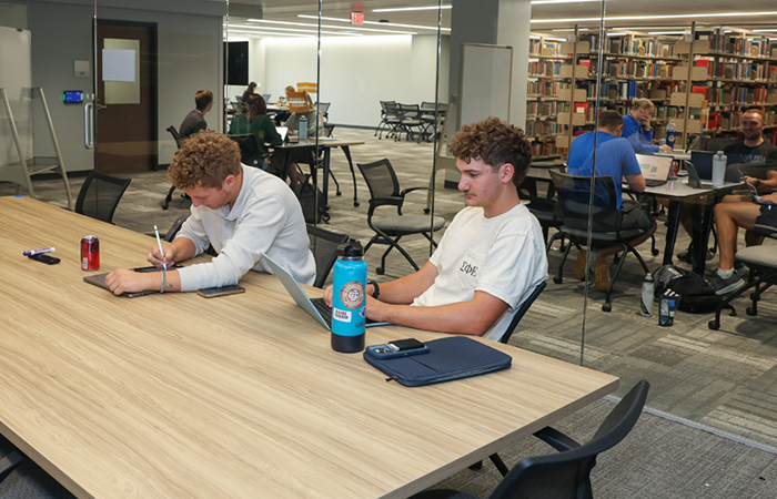 Two college-aged boys sit in an enclosed room with a glass wall at the library. One is writing on his tablet and the other has his laptop opened. Through the glass you can see the libraries stacks and two other tables with students gathered around them using their laptops.
