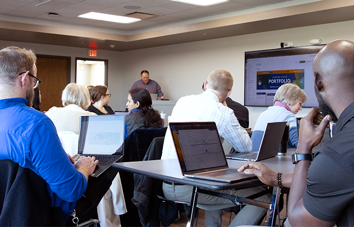 A group of people attending a presentation in a meeting room, focused on laptops with a projected slide reading "PORTFOLIO" visible in the background.