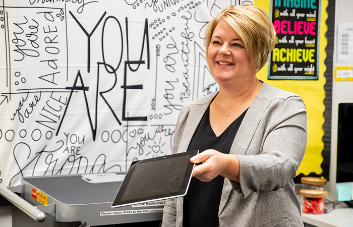 A woman hands out iPads in a bright classroom showcasing motivational posters.