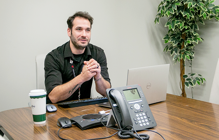 A man sitting at a conference table with a laptop, phone, and coffee cup, engaged in discussion.