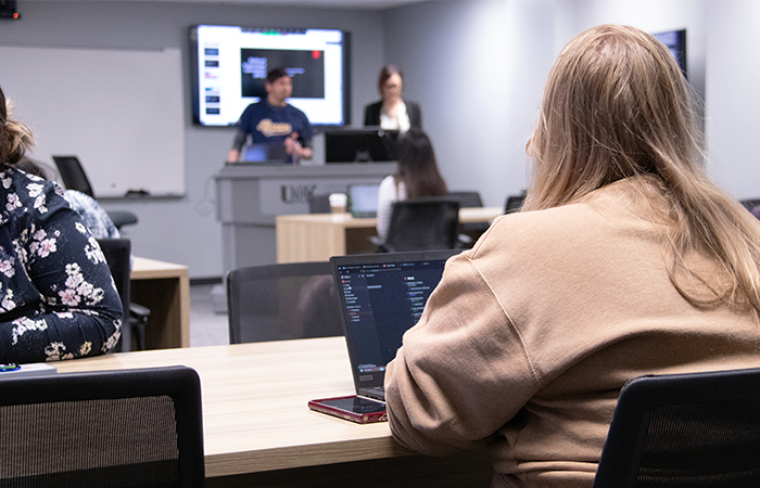 学生 engaged in a classroom setting, with two people presenting at the front and others using laptops.