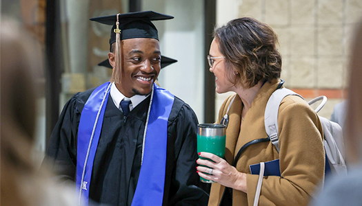 A graduate in a black cap and gown smiles at a woman holding a green tumbler