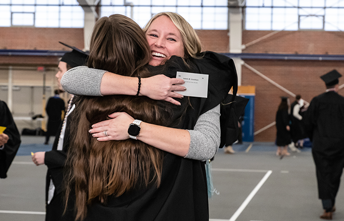 Two women embracing each other in a hug. The blonde woman is facing the camera and has a large smile on her face, the brunette woman's back is the camera and she's wearing a graduation robe.