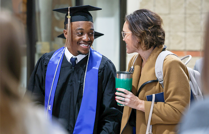 A graduate in a black cap and gown smiles at a woman holding a green tumbler.
