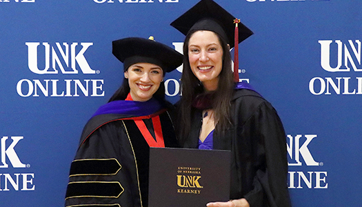 Two women celebrate at a graduation ceremony, one in academic regalia holding a diploma, with a backdrop featuring the 太阳城娱乐官网在线 logo.