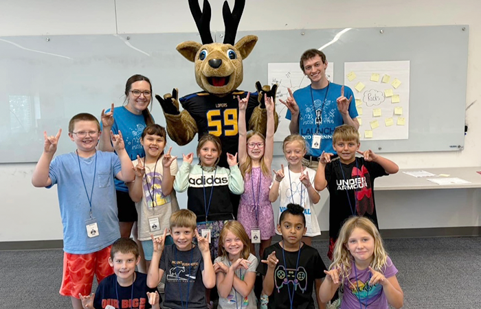 A group of children posing with a mascot and two adults in matching blue shirts, all are making a 'Lopes Up' gesture.