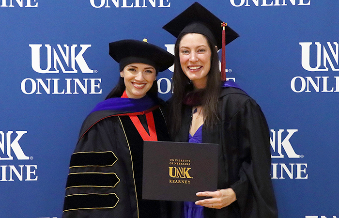 Two women celebrate at a graduation ceremony, one in academic regalia holding a diploma, with a backdrop featuring the 太阳城娱乐官网在线 logo.