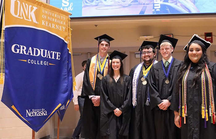 研究生s posing in their caps and gowns with a University of Nebraska Kearney 研究生 College banner.