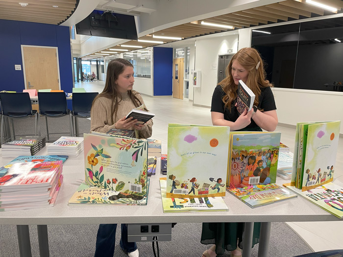 Two women are engaging in a discussion while browsing through a display of colorful books on a table.