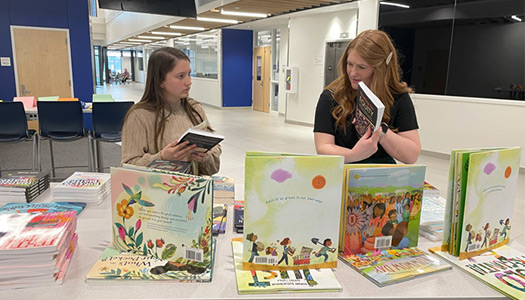 Two women are engaging in a discussion while browsing through a display of colorful books on a table.