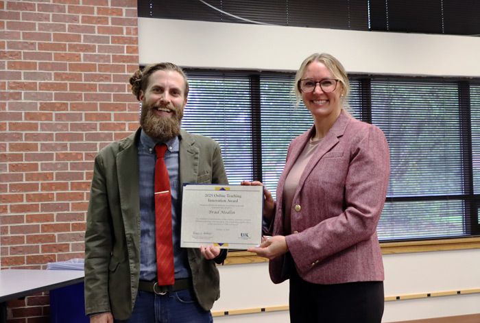 Two individuals smiling while holding a certificate; one is a man with a beard in a suit jacket and tie, and the other is a woman in a blazer.