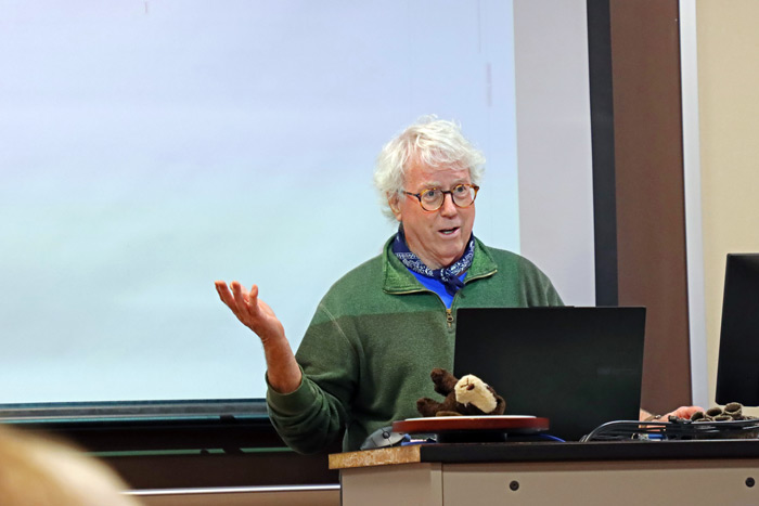 Rick Brown wearing glasses and a green sweater while gesturing with his hand in the front of a college classroom.