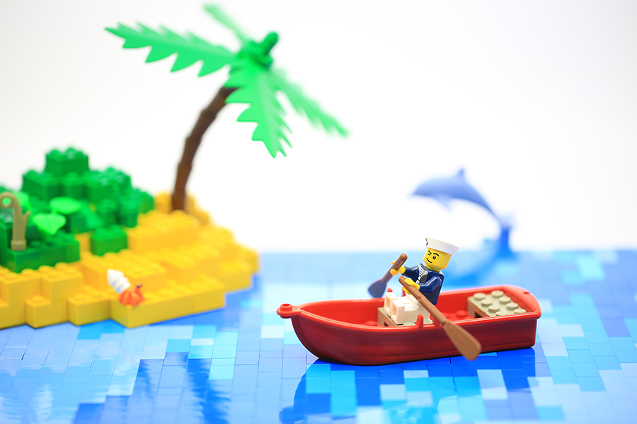 A playful LEGO scene featuring a sailor minifigure rowing a red boat near a tropical island with palm trees and a dolphin in the water.