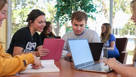 students sit at a table with ipads and laptops
