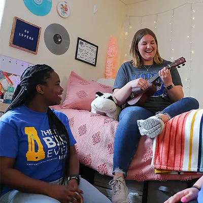a student plays a ukulele in their room while another student sits on the floor